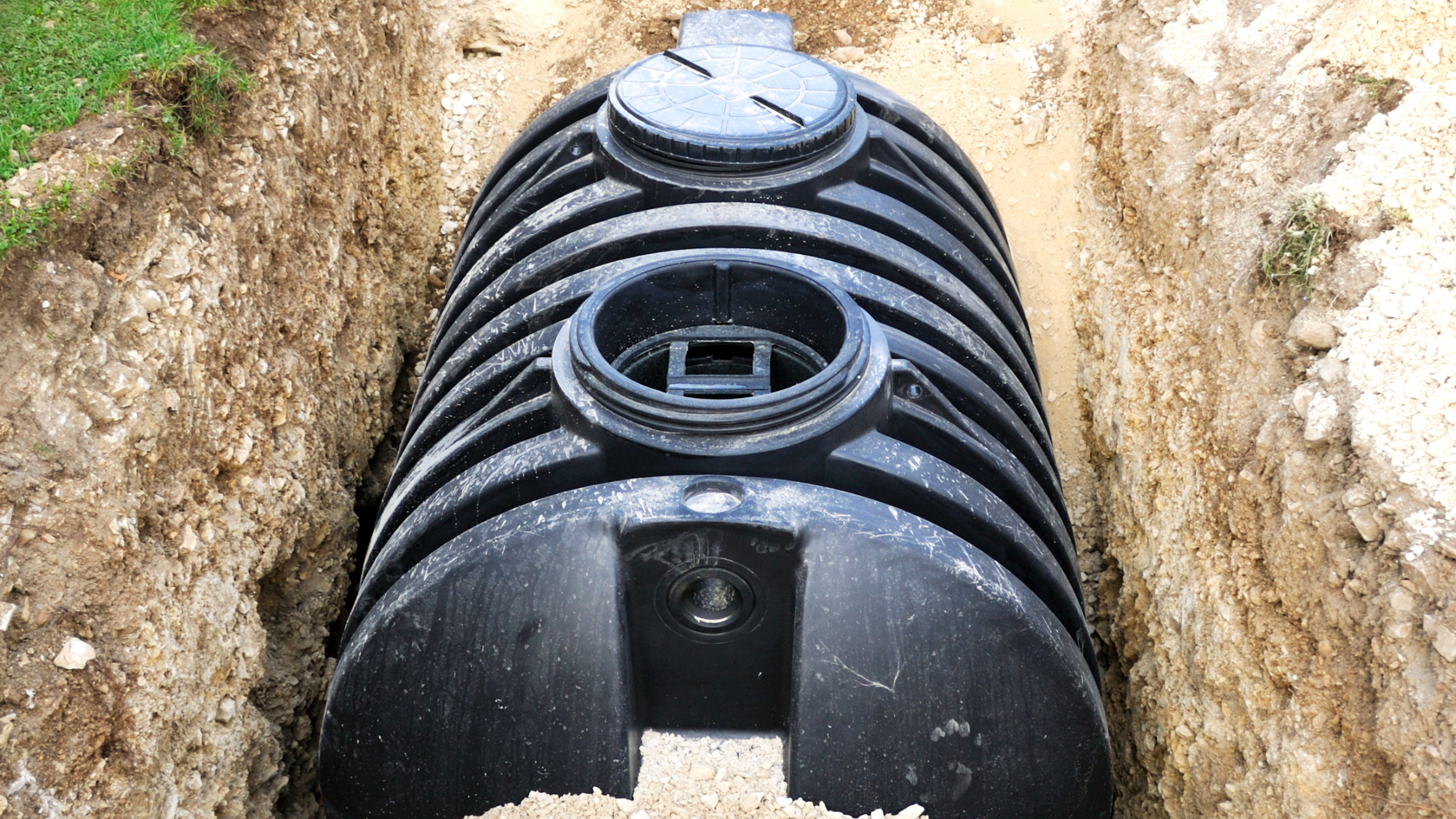 Black septic tank in hole surrounded by dirt and grass, presumably being installed. 